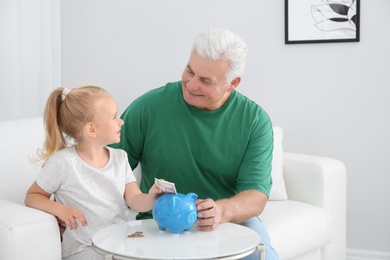 Photo of Little girl putting money into piggy bank and her grandfather at table