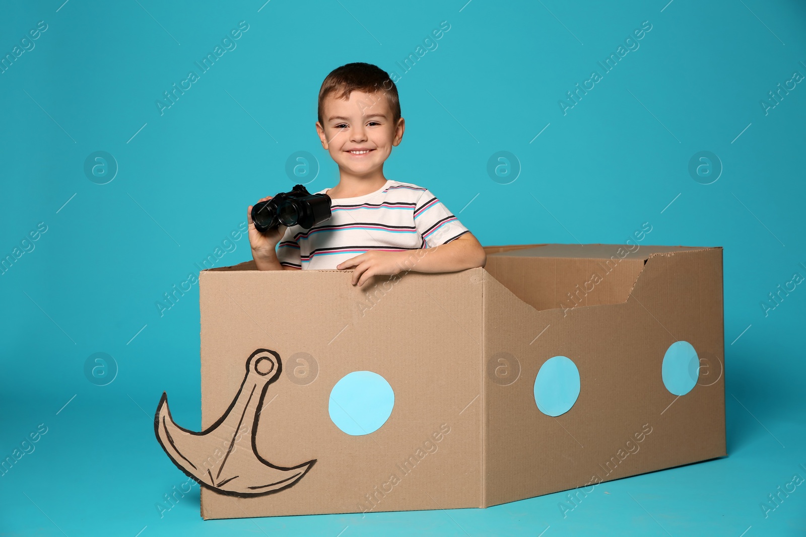 Photo of Cute little boy playing with binoculars and cardboard boat on color background
