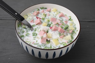 Photo of Delicious cold summer soup (okroshka) with boiled sausage and spoon in bowl on grey wooden table, closeup