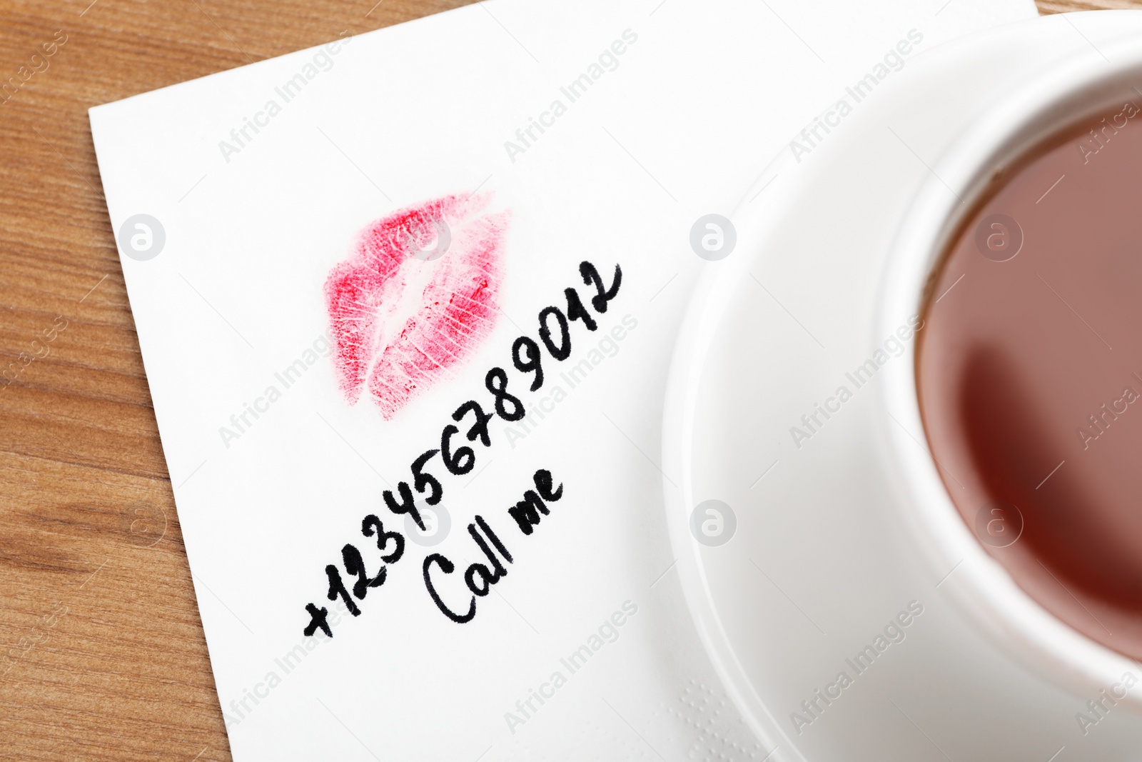 Photo of Paper napkin with phone number and lipstick mark under tea cup on wooden table, top view