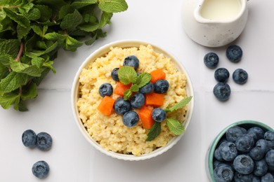 Tasty millet porridge with blueberries, pumpkin and mint in bowl on white tiled table, flat lay