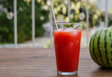 Photo of Glass of delicious watermelon drink with mint, ice cubes and fresh fruit on wooden table outdoors, space for text