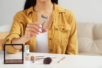 Woman with eyelash curler, makeup products and mirror at table indoors, closeup