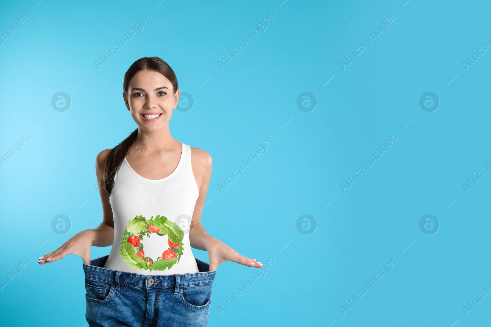 Image of Slim young woman wearing oversized jeans and images of vegetables on her belly against light blue background. Healthy eating