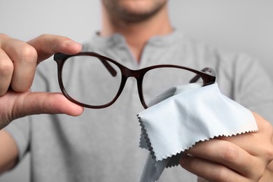 Man wiping glasses with microfiber cloth on light grey background, selective focus