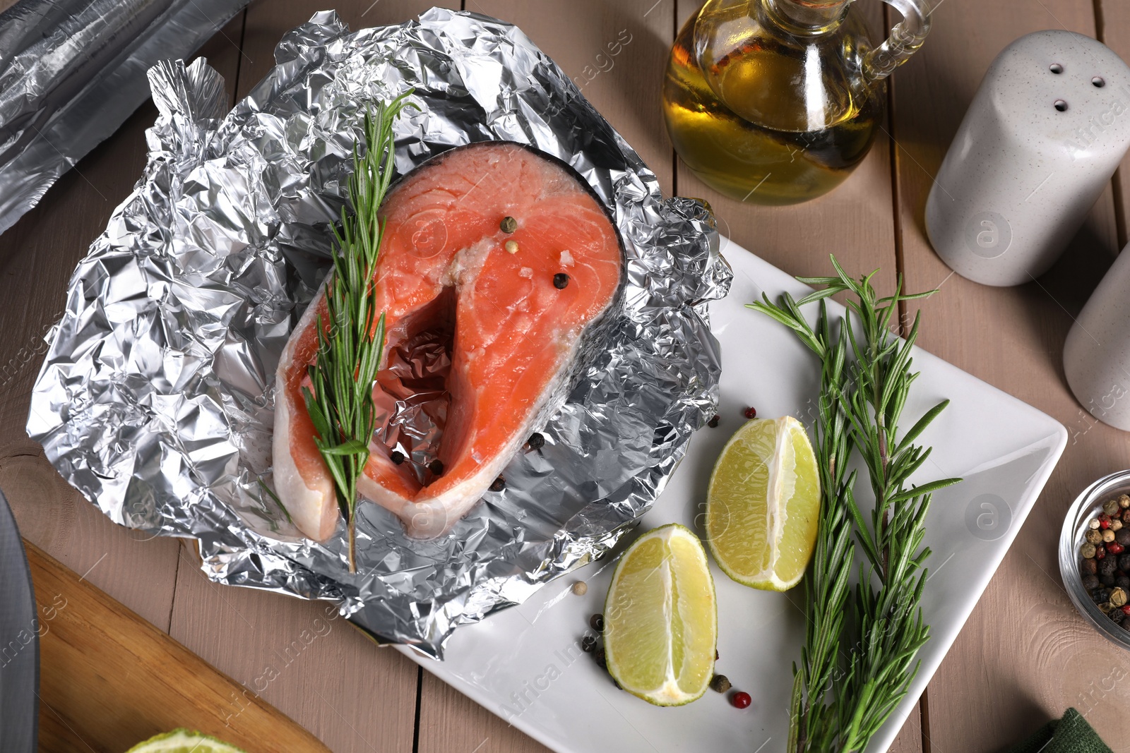 Photo of Aluminum foil with raw salmon, lime slices, rosemary and spices on wooden table, flat lay