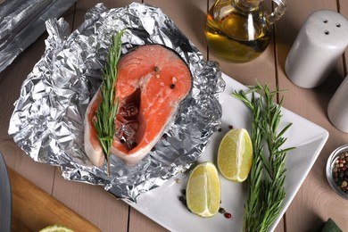 Photo of Aluminum foil with raw salmon, lime slices, rosemary and spices on wooden table, flat lay