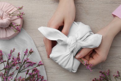 Photo of Furoshiki technique. Woman decorating gift wrapped in white fabric with beautiful pink flower at wooden table, top view