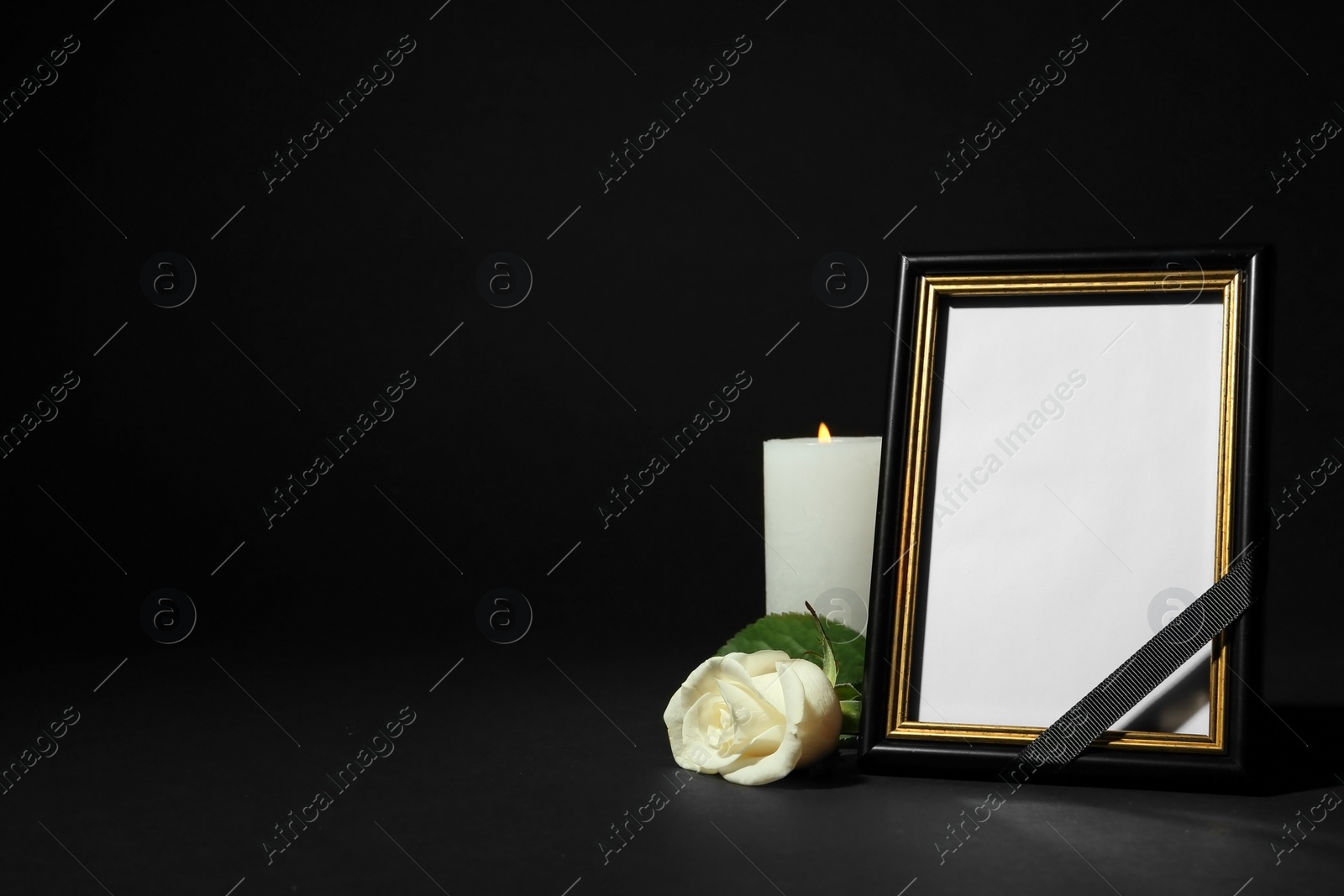Photo of Funeral photo frame with ribbon, white rose and candle on dark table against black background. Space for design