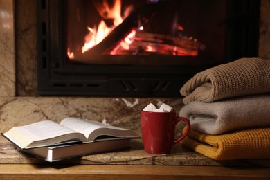 Stack of warm sweaters, cup, books and fireplace indoors
