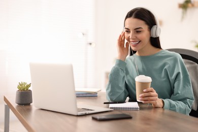 Photo of Young woman in headphones watching webinar at table in room