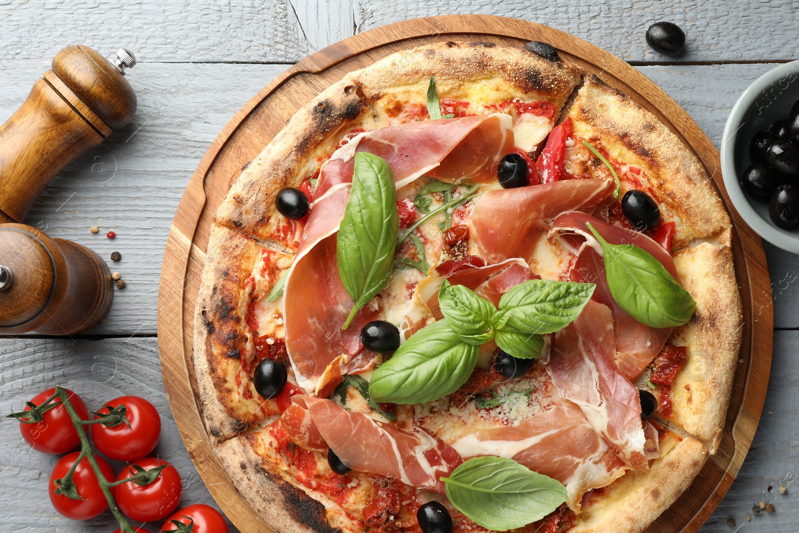 Photo of Tasty pizza with cured ham, olives, tomatoes and basil on gray wooden table, flat lay