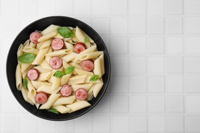 Photo of Tasty pasta with smoked sausage and basil in bowl on white tiled table, top view. Space for text