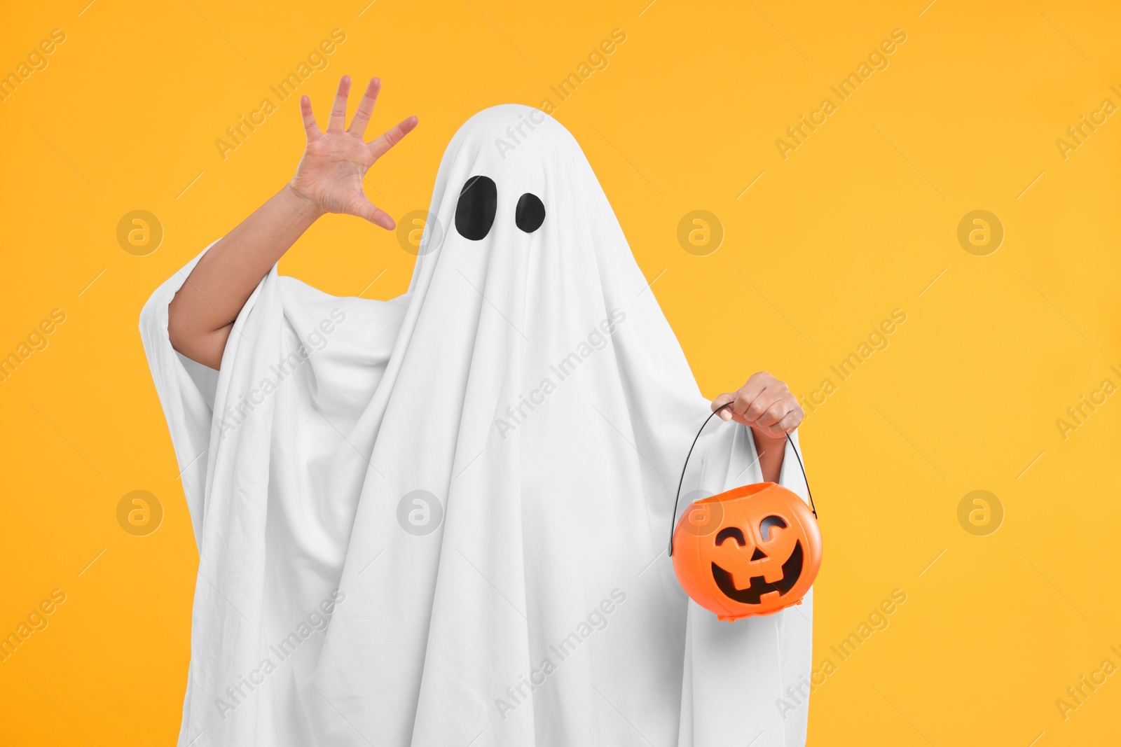 Photo of Woman in white ghost costume holding pumpkin bucket on yellow background. Halloween celebration