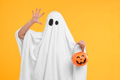Photo of Woman in white ghost costume holding pumpkin bucket on yellow background. Halloween celebration