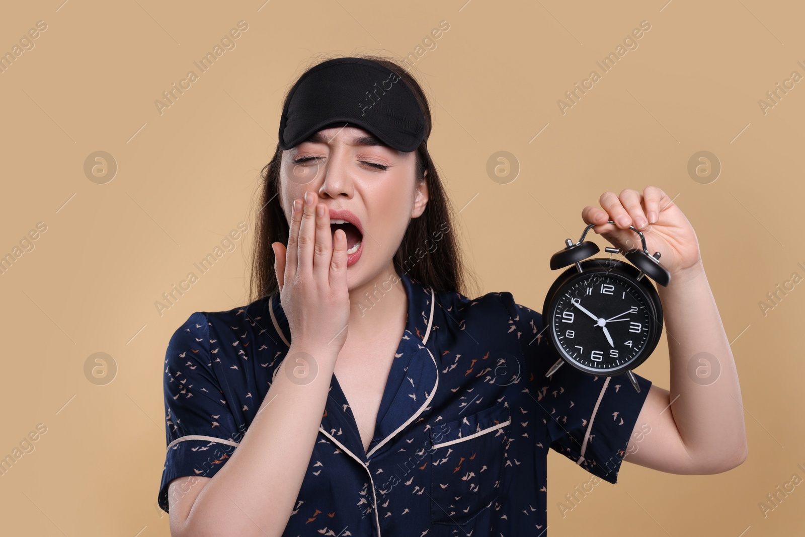 Photo of Tired young woman with sleep mask and alarm clock yawning on beige background. Insomnia problem