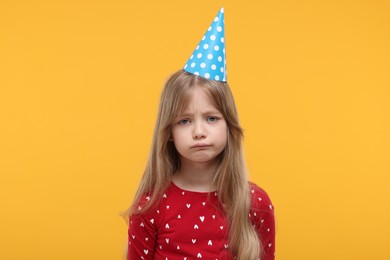 Photo of Unhappy little girl in party hat on yellow background