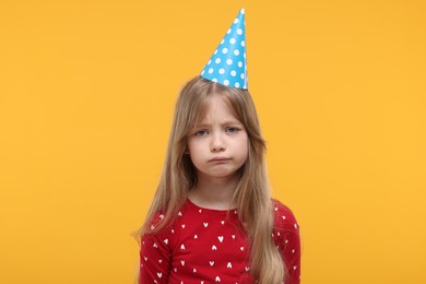 Unhappy little girl in party hat on yellow background