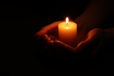 Woman holding burning candle in hands on black background, closeup