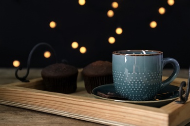 Photo of Cup with drink and piece of chocolate on wooden table against blurred lights, closeup. Space for text
