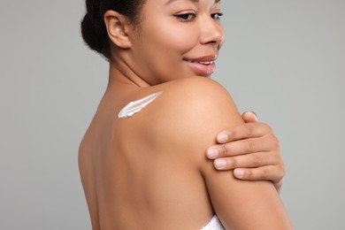 Young woman applying body cream onto back on grey background, closeup