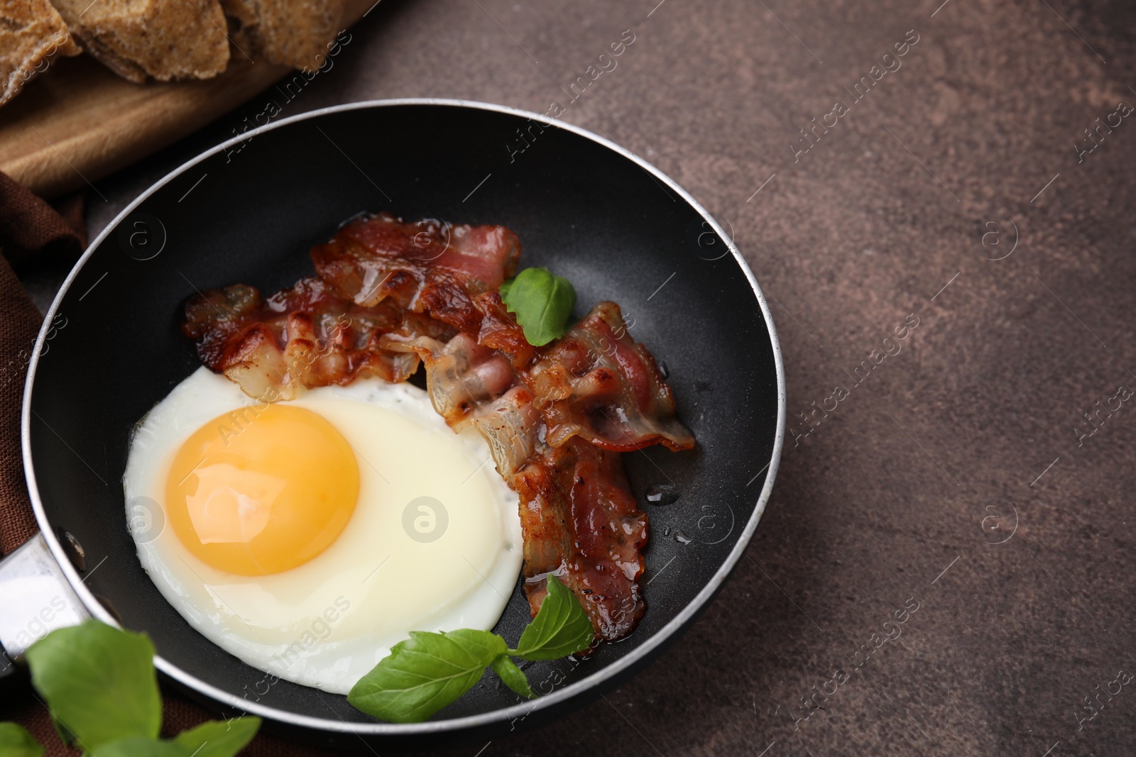 Photo of Fried egg, bacon and basil in frying pan on brown table, space for text
