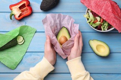 Photo of Woman packing half of fresh avocado into beeswax food wrap at light blue wooden table, top view