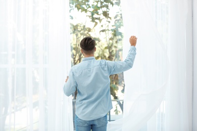 Photo of Young man opening window curtains at home