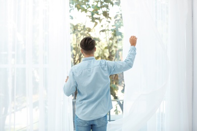 Young man opening window curtains at home