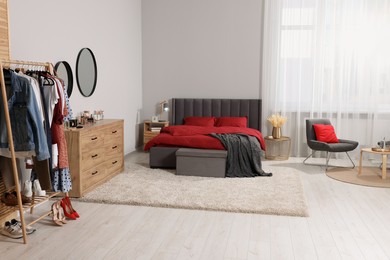 Photo of Stylish bedroom with comfortable bed, armchair, table, wooden chest of drawers and clothing rack. Interior design