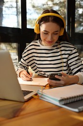 Young female student with laptop and headphones studying at table in cafe