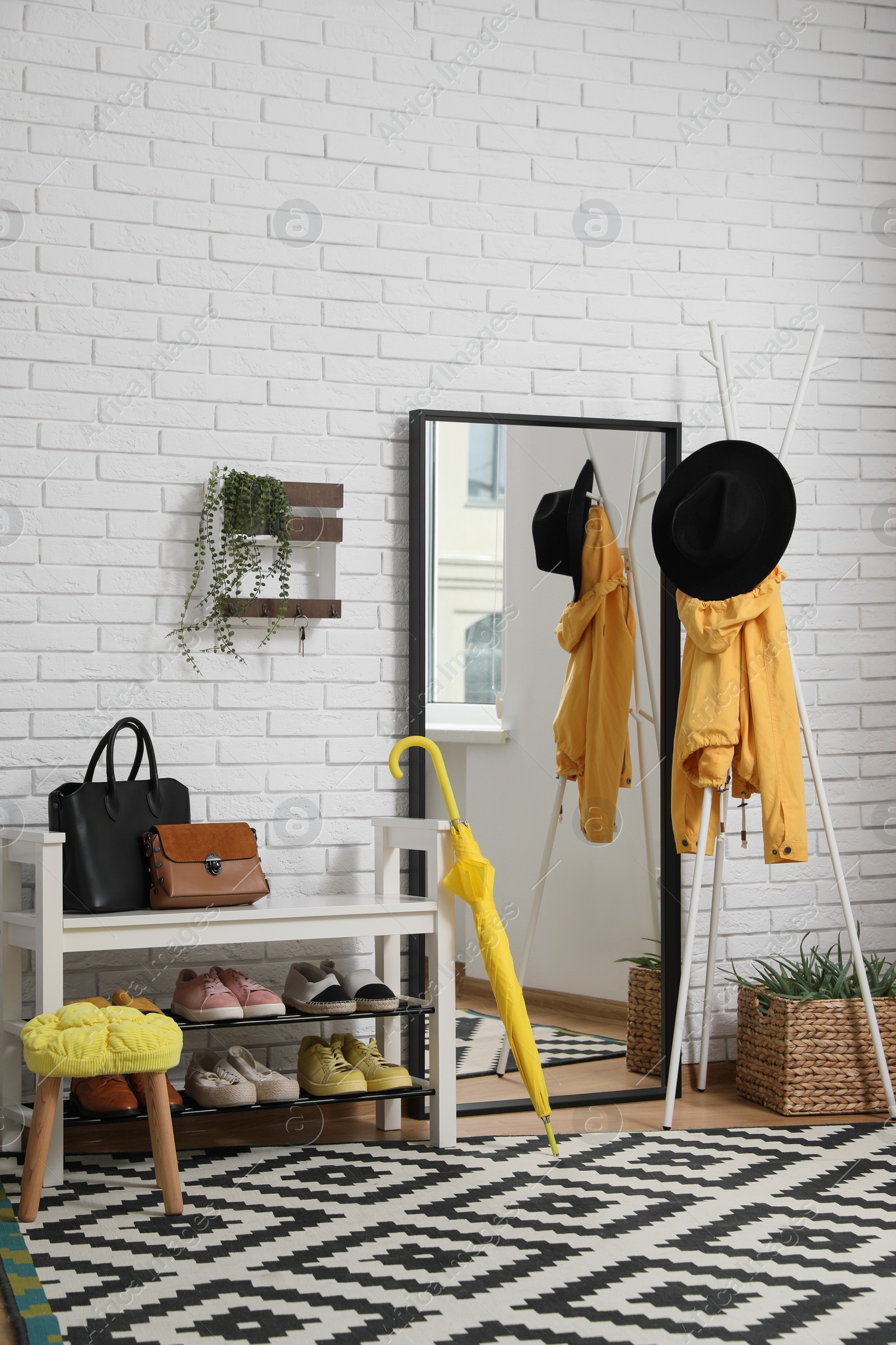 Photo of Stylish hallway interior with coat rack, shoe storage bench and mirror near white brick wall