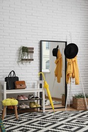 Photo of Stylish hallway interior with coat rack, shoe storage bench and mirror near white brick wall