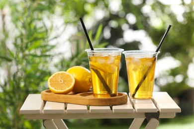 Photo of Plastic cups of tasty iced tea with lemon and fresh fruits on white wooden table against blurred green background