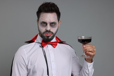 Photo of Man in scary vampire costume with fangs and glass of wine on light grey background. Halloween celebration