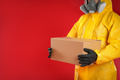 Photo of Man in chemical protective suit holding cardboard box on red background, closeup view with space for text. Prevention of virus spread