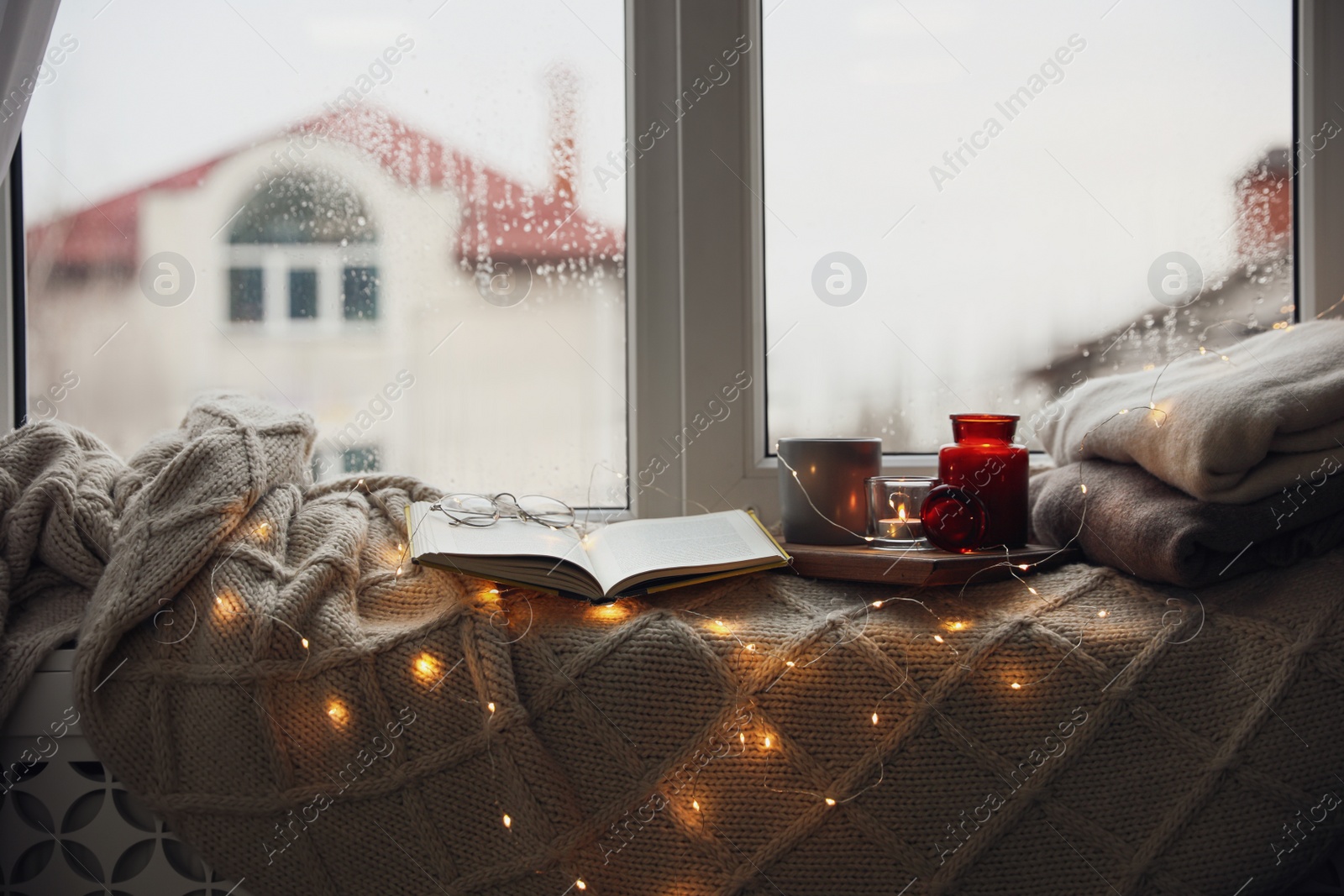 Photo of Cup of hot drink, open book and Christmas lights on knitted blanket near window. Cozy season