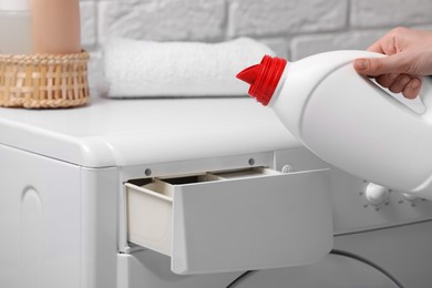Photo of Woman pouring fabric softener from bottle into washing machine near white brick wall, closeup