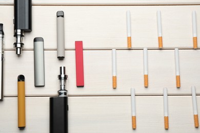 Different vaping devices and cigarettes on white wooden table, flat lay. Smoking alternative