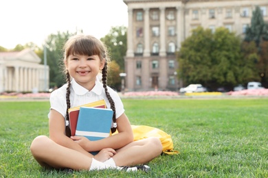 Cute girl with school stationery sitting on green lawn outdoors
