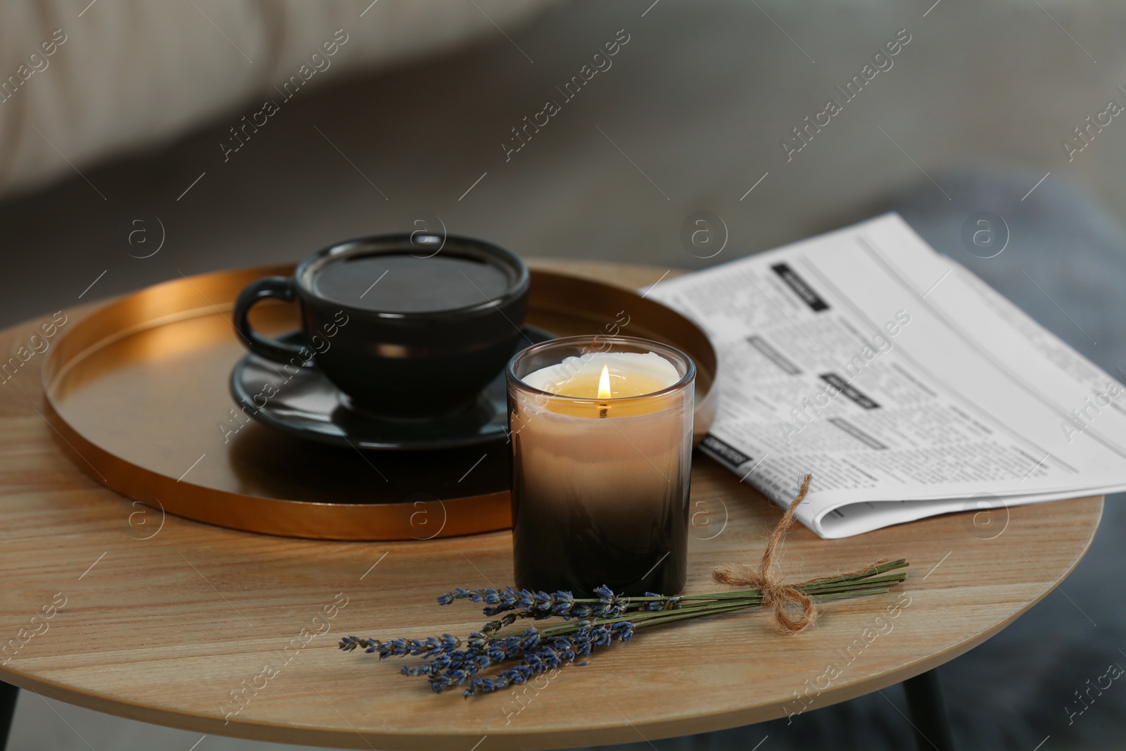 Photo of Beautiful candle, lavender, newspaper and cup of coffee on round wooden table indoors