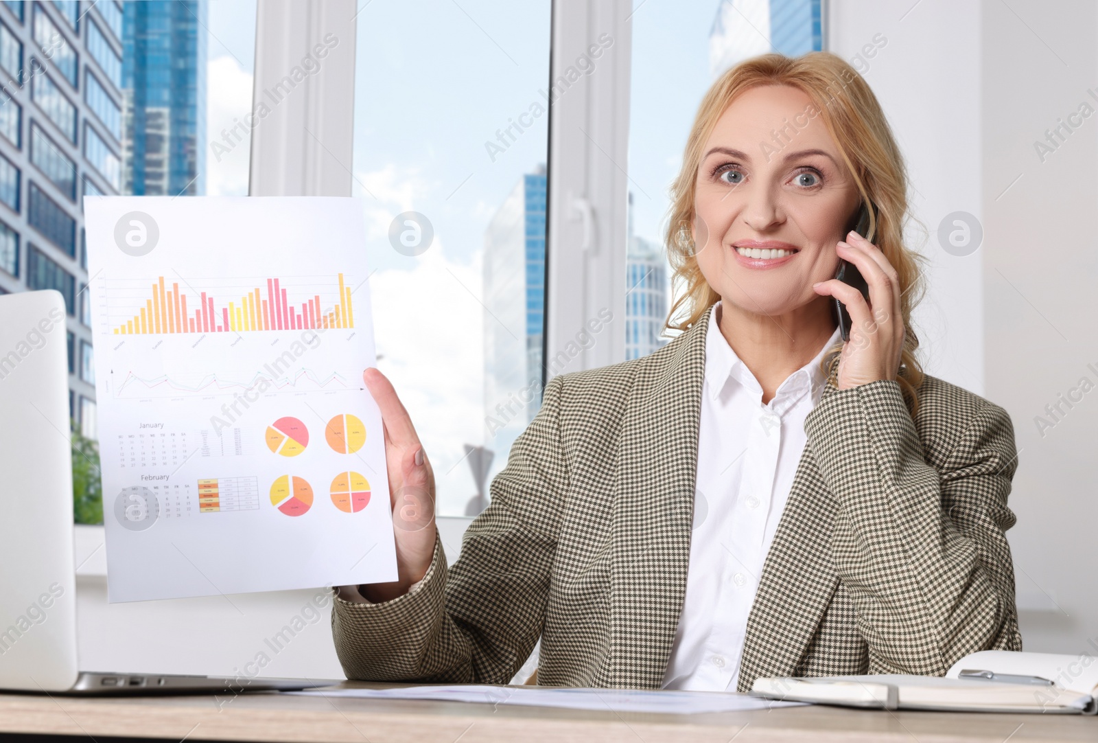 Photo of Lady boss talking on smartphone and showing papers with statistics at desk in office. Successful businesswoman