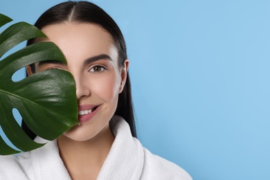 Photo of Woman in bathrobe holding leaf of monstera on light blue background, space for text. Spa treatment