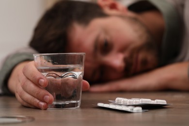 Photo of Depressed man with glass of water and antidepressant pills at table, selective focus