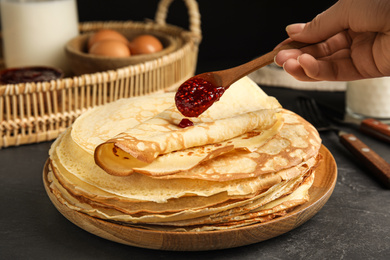 Photo of Woman putting jam on stack of fresh thin pancakes at black table, closeup