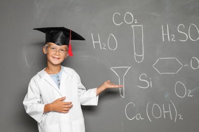 Little school child in laboratory uniform with graduate cap and chemical formulas on grey background