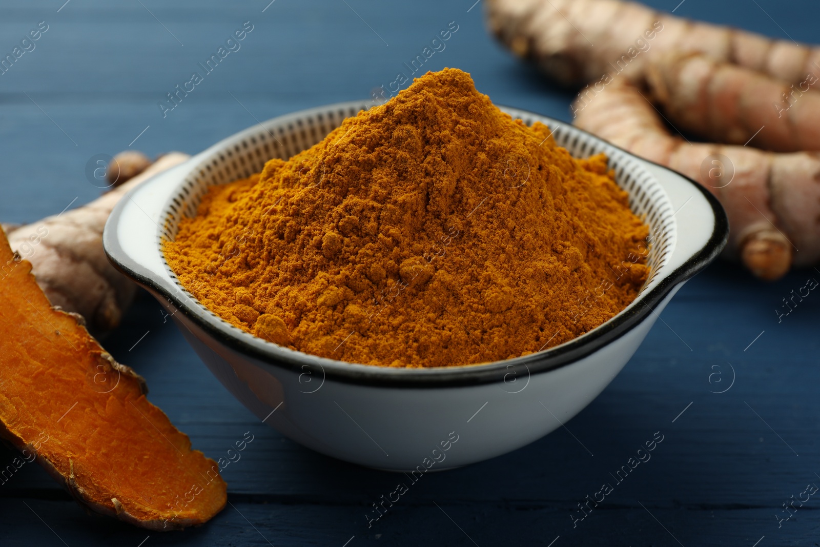 Photo of Bowl with turmeric powder and raw roots on blue wooden table, closeup