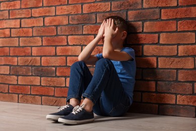 Photo of Child abuse. Upset boy sitting on floor near brick wall indoors, space for text