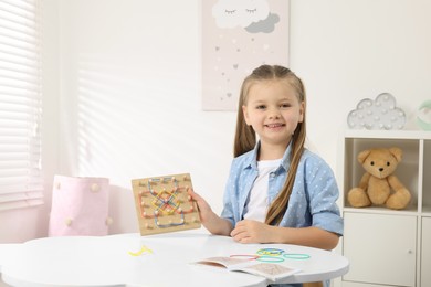 Photo of Motor skills development. Smiling girl showing geoboard and rubber bands at white table in room. Space for text