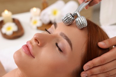 Photo of Young woman receiving facial massage with metal roller in beauty salon, closeup